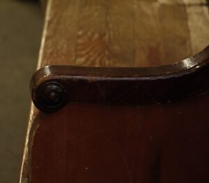 Close-up of an oak pew armrest and seat end in dark wood tone, showing the rounded support detail and worn surface.