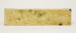 Single ceramic tile with a shiny crackled glaze in yellow tones and subtle green and purple mottling, shown in a close-up that reveals minor edge chips and age-related wear.