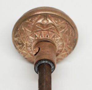 Close-up of a brass door knob backplate with an ornate fourfold pattern and a spindle extending downward.