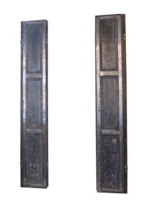 Pair of steel bi-fold shutters shown upright side by side, with three recessed panels on each section and cast iron trim details visible.