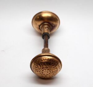 Pair of polished bronze door knobs with a spindle, shown in close-up with one plain knob and one patterned knob face.