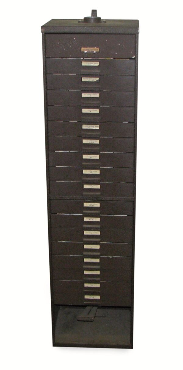 Tall narrow brown metal cabinet with many labeled drawers, shown front on with a foot press at the bottom.
