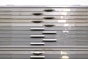 Front view of a 20-drawer steel flat file cabinet with mixed handles, visible wear, rust, and labels, showing the stacked drawers and the overall restoration-needed condition.