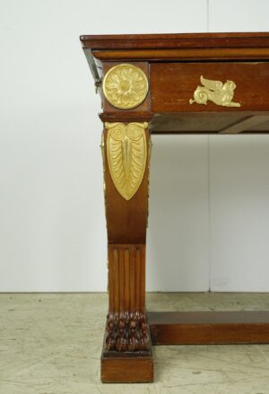 Close-up of an Empire-style desk corner with a mahogany leg, gilt bronze floral and winged appliques, and carved foot detail.