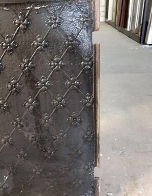 Close-up of a black cast iron fire back panel with raised fleur-de-lis detailing and a worn edge.