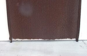 Wrought iron elevator gate with a solid lower panel, shown in close-up with visible deterioration along the bottom edge.