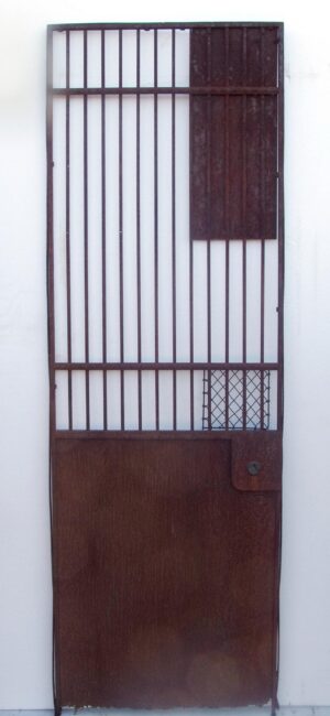 Wrought iron elevator gate with vertical bars, a solid lower panel, and a small mesh section near the bottom right, shown frontally with the original knob visible.