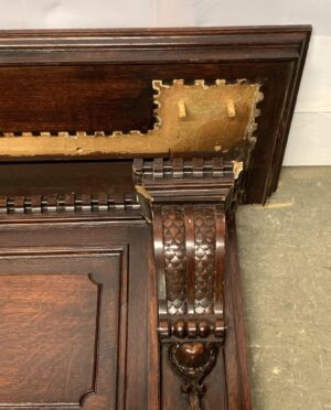 Close-up of a walnut fireplace mantel in dark wood tone, showing carved Rococo details and a reclaimed section with exposed backing and two small square hooks.