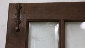 Close-up of a dark wood tone French door showing the upper section, with a metal hinge and beveled glass panes divided by wooden muntins.
