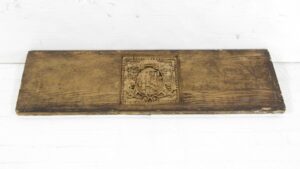 Brown Arts & Crafts pediment with a centered coat of arms carving, shown horizontally with worn edges and surface wear.