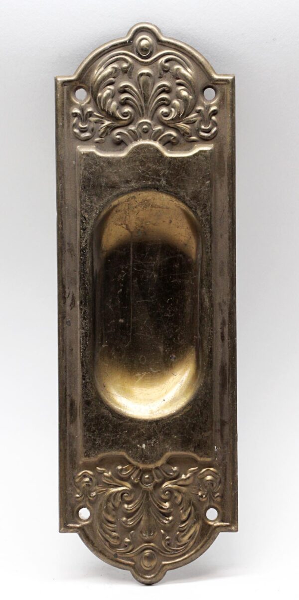 Decorative recessed pocket door plate with an antique brass finish and ornate embossed detailing, shown with four mounting holes and a central pull recess.