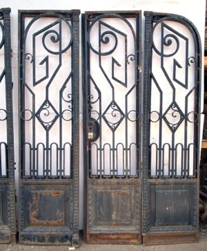 Art Nouveau wrought iron bifold doors with arched tops, scrollwork panels, and plain lower panels showing wear and rust.