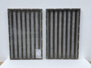 Pair of bronze window grates with woven brass mesh screens, shown side by side, with a small cutout in the lower corner of one grate.