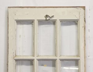 Vintage white 18-lite French door with six visible glass panes and worn painted wood trim, shown close up from the upper section with a hook latch at the top.
