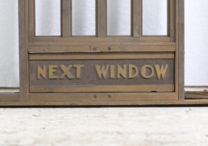 Bronze bank teller door with Art Deco lettering reading “Next Window” on the lower panel.
