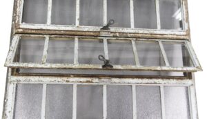 Large industrial encasement window with a heavy steel frame, showing multiple hinged louver-style panes and textured glass, with visible rust and wear on the frame.