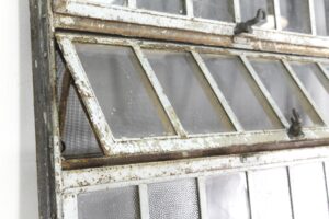 Close-up of an industrial encasement window showing hinged louver-style panes in a heavy steel frame with chipped finish and visible rust.