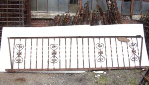 Heavily rusted wrought iron fire escape balcony front with ornate scroll details and a bent frame, shown leaning against a white backing board.