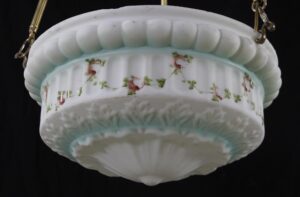 Close-up of a cast glass pendant light with a white dish shape, painted pink floral and green foliate details, blue accents, and brass suspension poles visible at the edges.