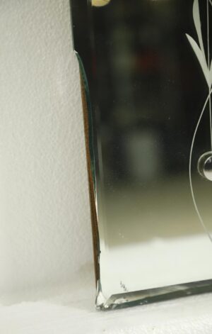Close-up of a beveled wall mirror with etched floral details, showing a broken corner and chipped edge on the mounted chipboard backing.