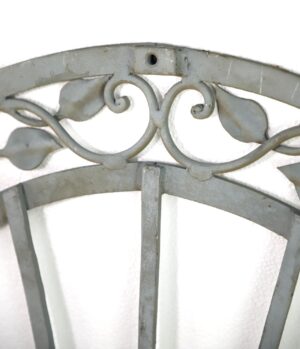 Gray wrought iron wall accent with arched top, curled vine detail, and leaf shapes, shown in close-up with some rust visible.