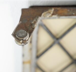 Close-up of a reclaimed steel casement window corner with rust, chipped paint, and a cracked leaded glass pane in the background.
