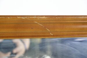 Close-up of a rectangular wall mirror frame in medium wood tone with visible surface wear and scratches near the edge.