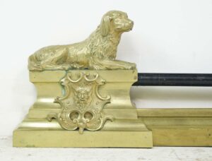 Antique brass fireplace fender with a reclining spaniel dog finial and ornate central crest detail.