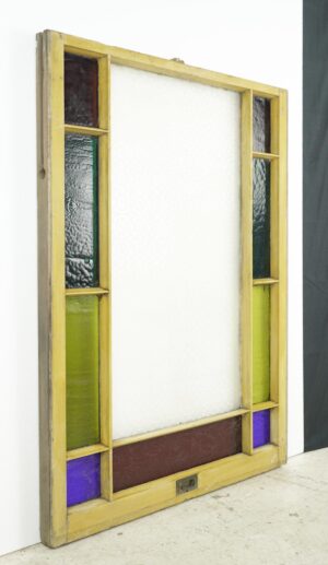 Antique pine framed Queen Anne window with clear center glass and narrow stained glass panels in green, purple, red, and dark tones around the edges, shown upright with visible wear on the frame.