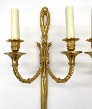 Close-up of a gilt bronze wall sconce with two curved arms, ribbon-tied center stem, and tapered candle sleeves, shown on a plain background.