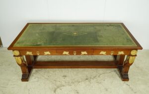 French Empire gentleman’s desk in dark wood with a green leather top, gilt bronze appliques, three front drawers with keyholes, and carved legs with gold accents.