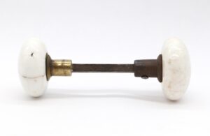Pair of white ceramic door knobs with a spindle, shown with one cast iron shank and one brass-plated steel shank.