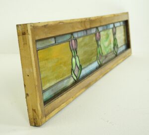 Art Nouveau stained glass window in a light wood pine frame, shown at an angle with leaded panes in multiple colors and a visible worn surface.