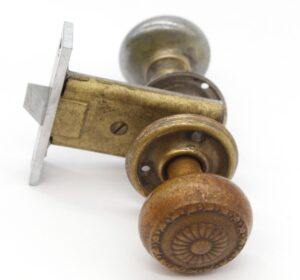 Close-up of a passage door knob set with one brass patina knob and rosette and one nickel patina knob and rosette, shown with the mortise latch and strike plate.