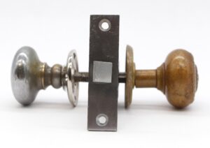 Passage door knob set with one brass-patina knob and one nickel-plated knob, shown with matching rosettes, a mortise, a strike plate, and a spindle.