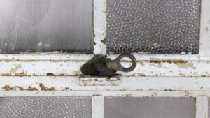 Close-up of an industrial encasement window showing a central metal latch on a heavy steel frame with textured glass panes and visible rust and peeling paint.