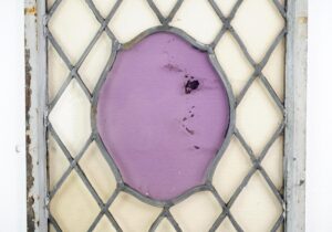 Steel framed casement window with crisscross leaded glass and a central purple shield-shaped panel, shown close up with chipped white paint and visible wear.