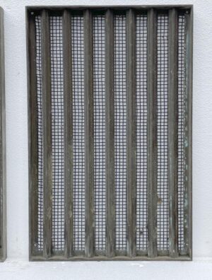 Antique bronze window grate with vertical bars over a woven brass mesh screen, shown front-facing with the lower corner cutout visible.