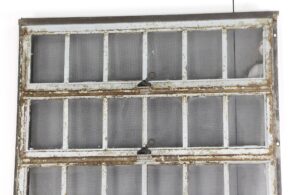Industrial encasement window with a heavy steel frame, divided into multiple panes with screened glass and two visible latch handles, shown with rust and worn paint on the frame.