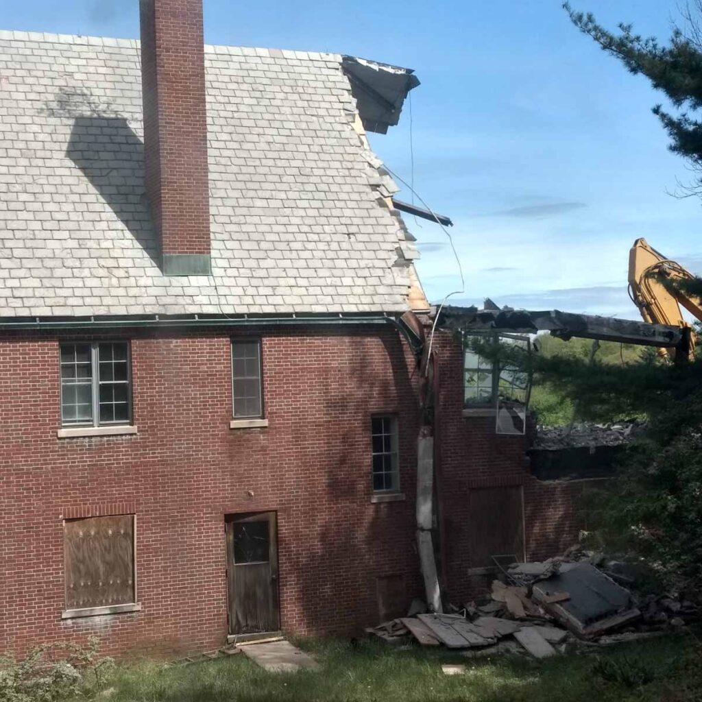 An excavator tears down a brick house near Mount Kisco, NY; roof and walls partially collapsed, debris scattered across the lot similar to Rose Hill Mansion’s grounds.