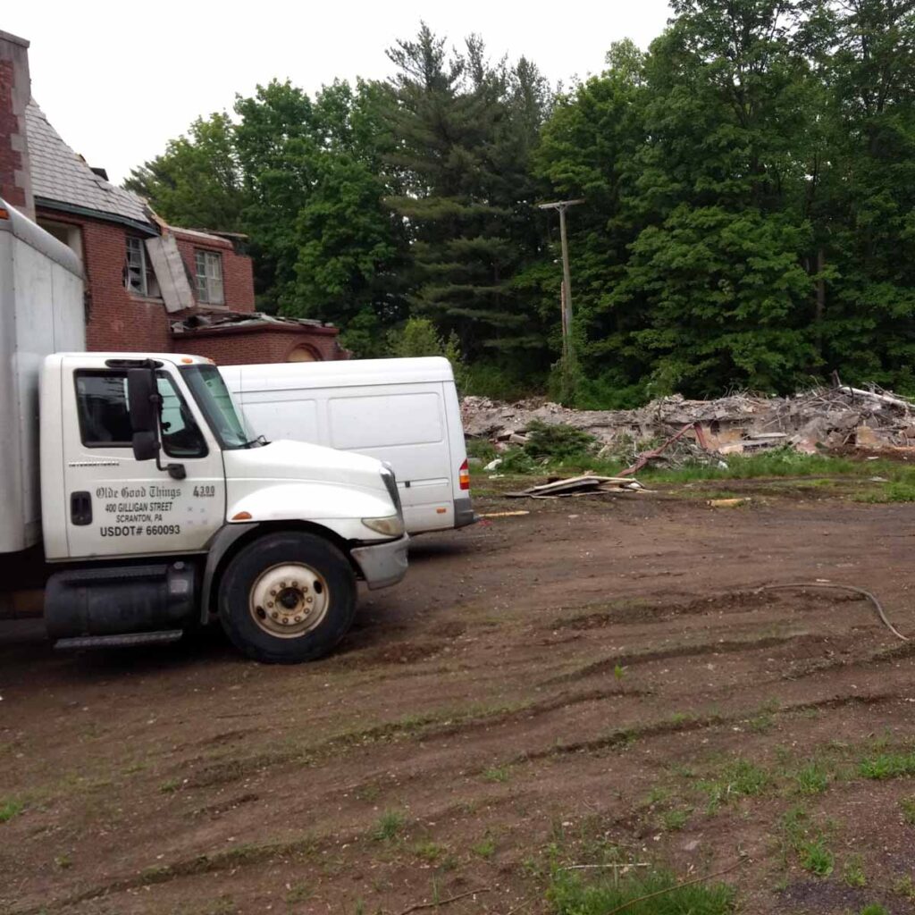 Two trucks stand on dirt beside a partly demolished brick building in Mount Kisco, NY. Debris and rubble cover the ground, with trees surrounding the area in the background.