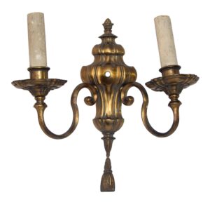 Traditional bronze two-arm wall sconce with urn detail, curved arms, and a tassel finial, shown with two candle holders.