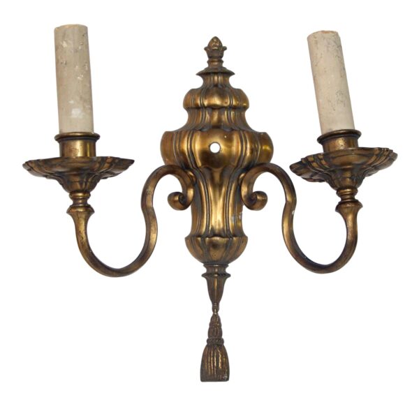 Traditional bronze two-arm wall sconce with urn detail, curved arms, and a tassel finial, shown with two candle holders.