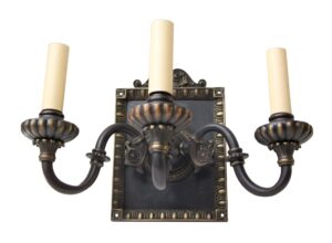 Tudor-style metal wall sconce with three candle-like lights and a decorative black and gold backplate, shown front on.