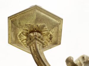 Close-up of an antique brass wall sconce detail with a hexagonal backplate and curved arm, showing worn surface and floral ornamentation.