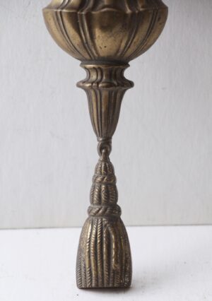 Close-up of an antique bronze wall sconce showing the urn-shaped top, fluted stem, and tassel detail.