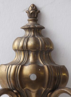 Close-up of an antique bronze wall sconce with a tassel finial and urn detail, shown from the front with one mounting hole visible.