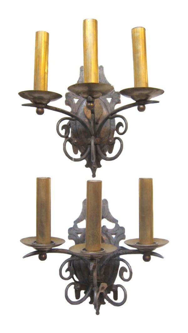Pair of brass three-arm wall sconces with a black finish, shown as two matching fixtures with upward candle-style holders and ornate scrollwork backplates.