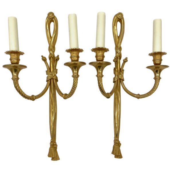 Pair of gilded bronze wall sconces with two candle arms each, ribbon tassel details, and cream candle sleeves, shown as a matching pair.