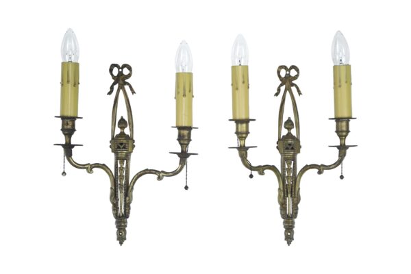 Pair of neoclassical brass wall sconces with two candle-style arms, ornate foliate detailing, and dangling chain pulls, shown as a matched pair.